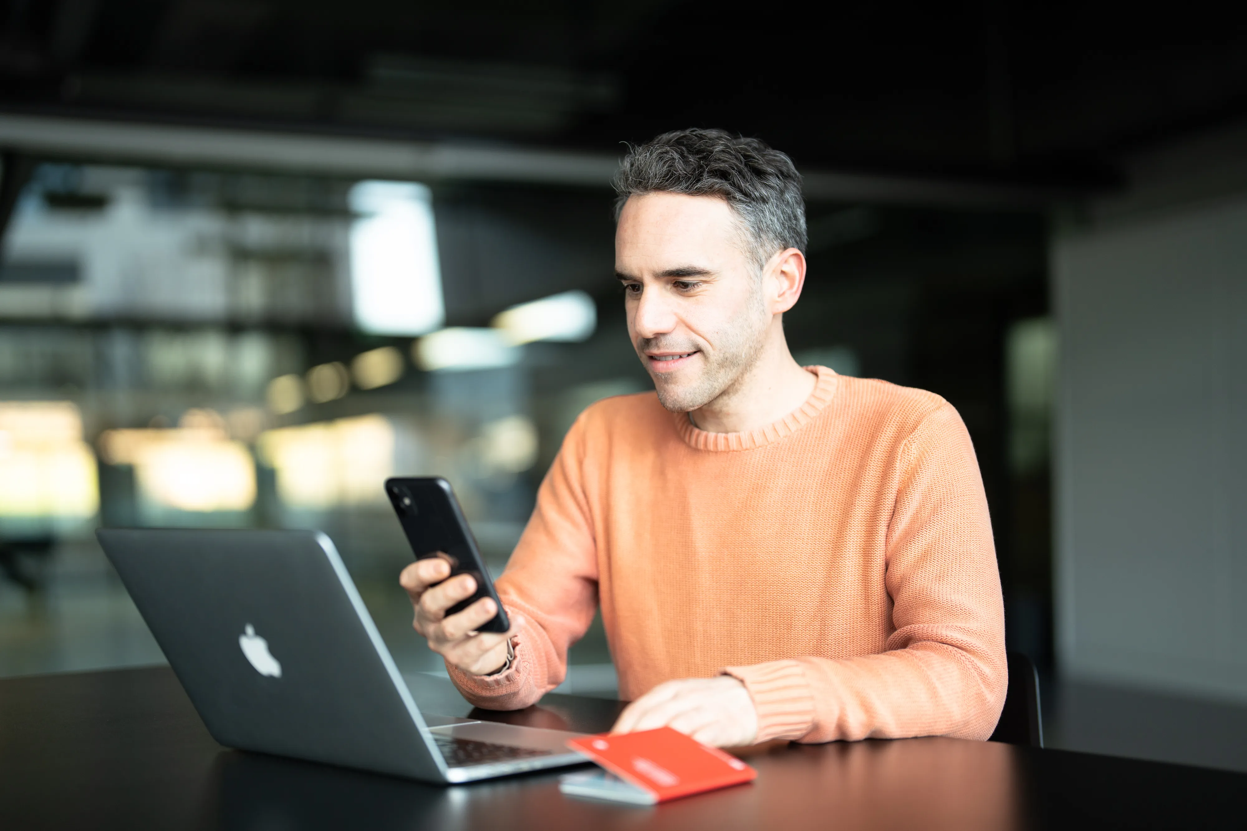 ein Mann sitzt vor dem Laptop und hält sein Mobiltelefon davor. Neben ihm liegt ein Schweizer Pass